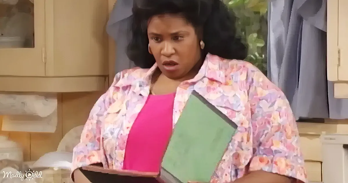 A woman in a floral shirt holds a green and pink book in a kitchen setting.
