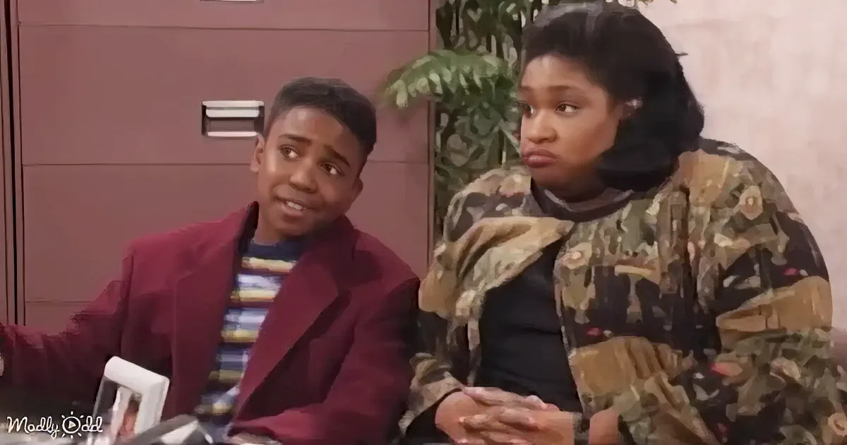 A young man in a maroon blazer and striped shirt sits next to a woman in a camouflage jacket, both appearing surprised or...