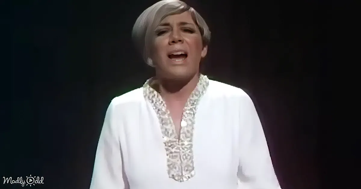 A person in a white, ornate top is singing on stage with a dark background.