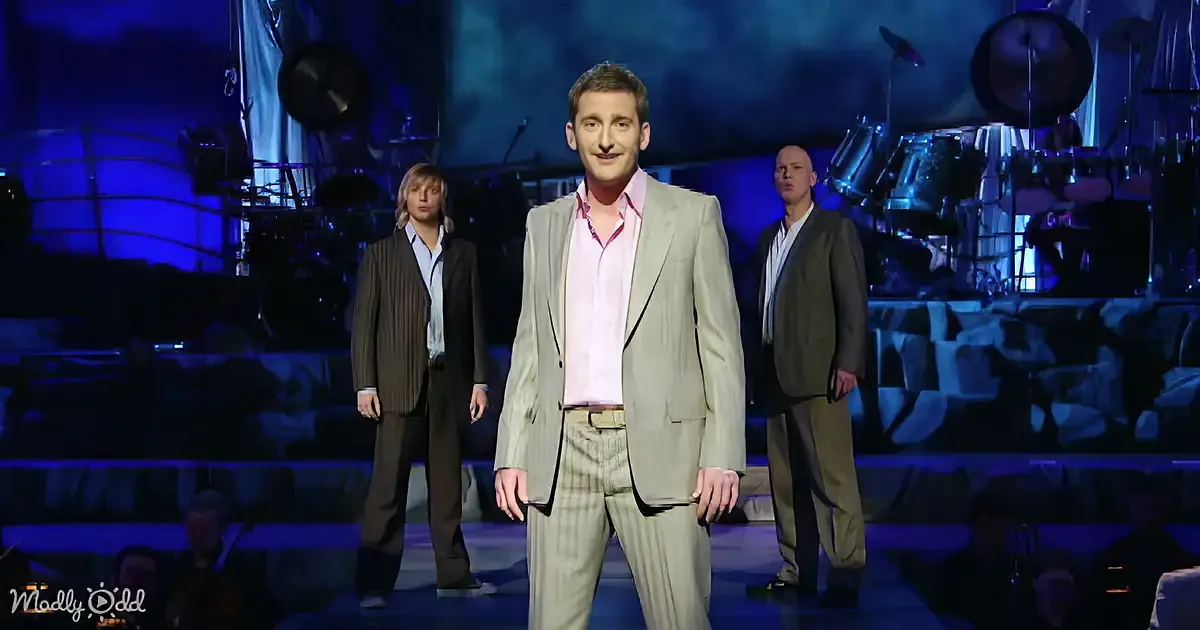 A man in a beige suit stands center stage with two others, a band setup behind, under blue stage lighting.
