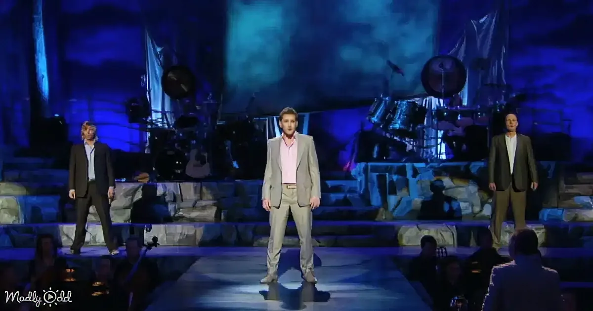 A man in a light suit stands center stage, flanked by two others, with a band and blue lighting in the background.