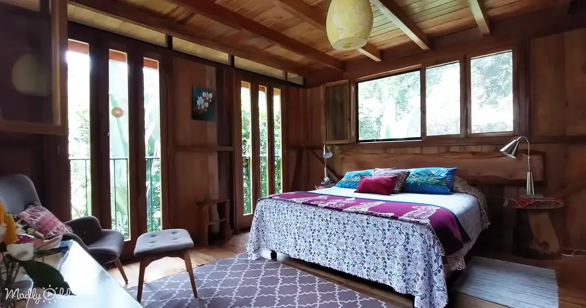 A cozy wooden bedroom with a bed, colorful pillows, and large windows overlooking greenery.