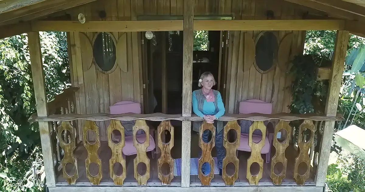 A woman sits on a wooden balcony with a unique railing, surrounded by greenery.