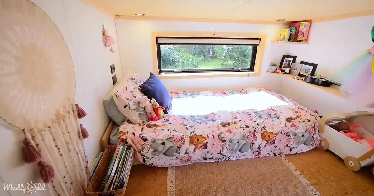 A cozy bedroom with a floral bedspread, a window showing greenery, and shelves with books and decor.