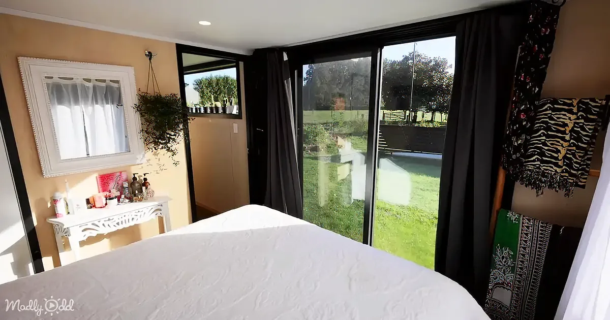 A cozy bedroom with a white bed, a dresser, a mirror, and a large sliding glass door leading to a garden.