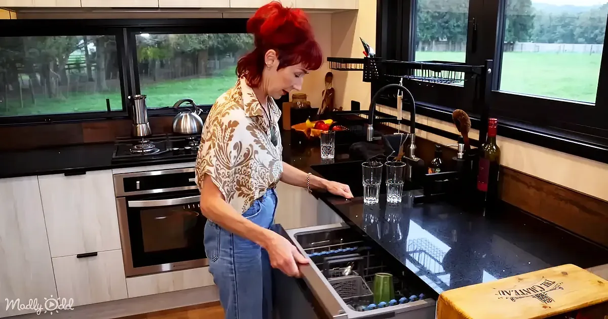 A woman with red hair stands in a modern kitchen, opening a dishwasher.