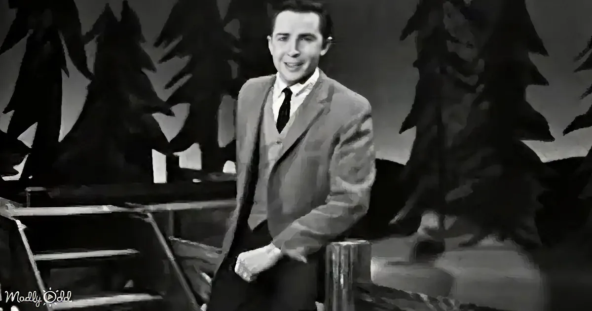 A man in a suit sits on a stool, smiling, with a backdrop of trees and a stage setting.