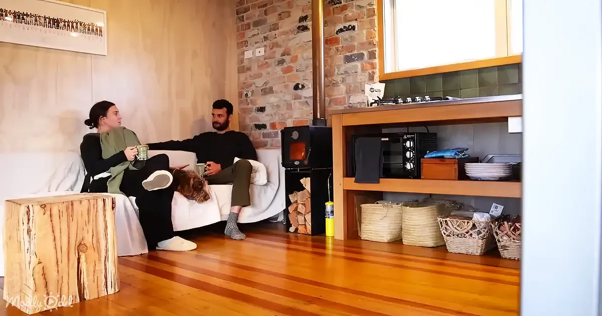 A couple sits on a couch, holding mugs, in a cozy room with a brick wall, wood stove, and wooden floor.