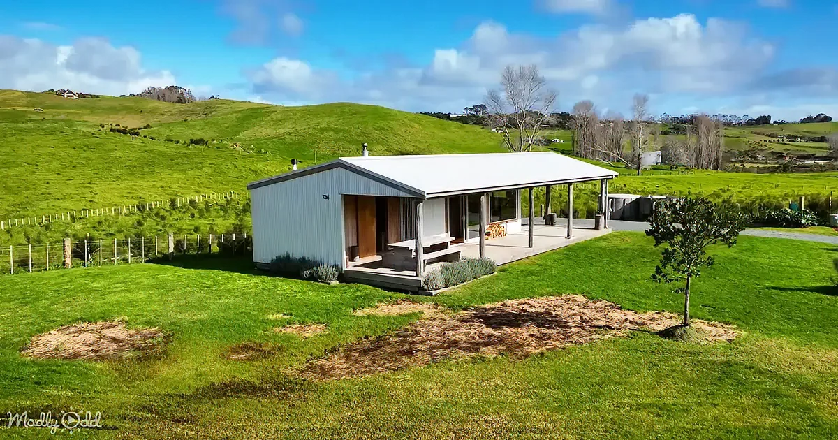 A modern, single-story house with a porch sits amidst lush green fields under a partly cloudy sky.