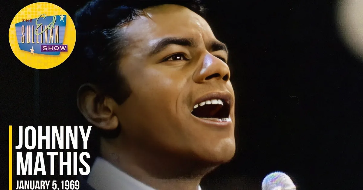 A man in a suit sings into a microphone, his face illuminated against a dark background.