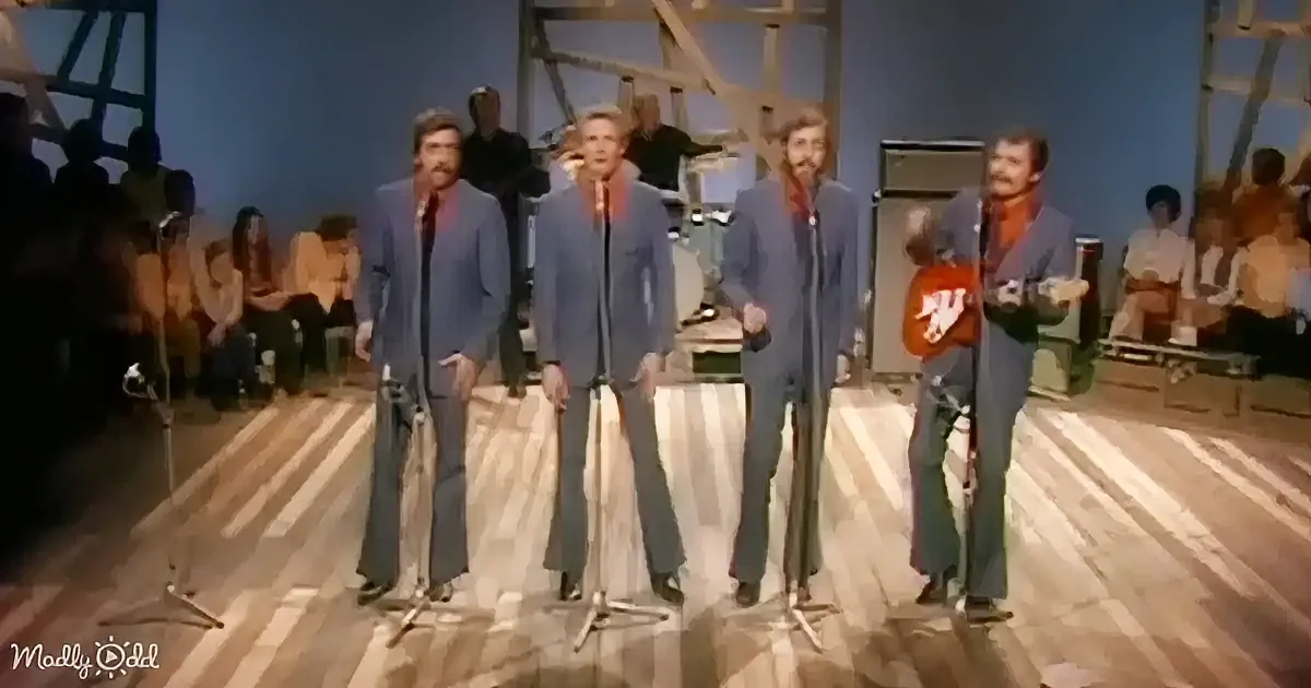 Four men in blue suits sing on stage, one playing a guitar, with an audience and stage equipment in the background.