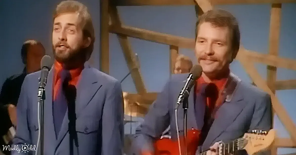 Two men in suits sing into microphones, one playing guitar, against a wooden backdrop.