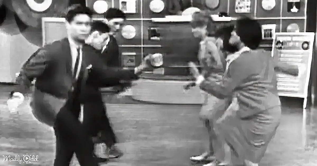 A man in a suit holds a gun, aiming at a group of people in a room with clocks and shelves.