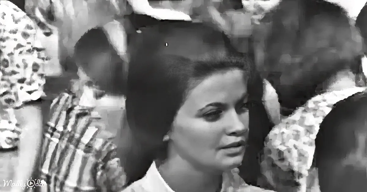A woman with dark hair and a white shirt stands in a crowd, looking to the side.