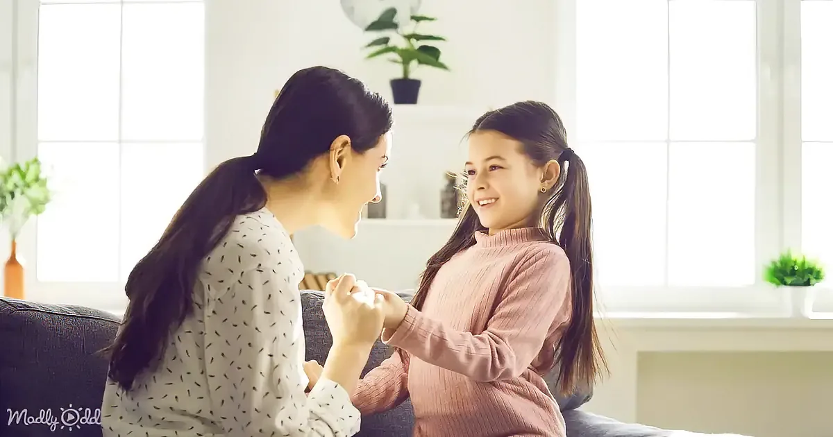 A woman and a girl are sitting on a couch, holding hands and smiling at each other in a bright, cozy room.
