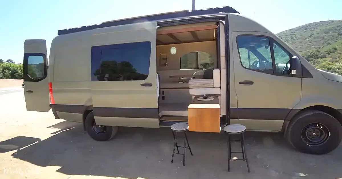 A beige van with its side door open, revealing a cozy interior setup with a chair, table, and stools, parked on a sunny day.