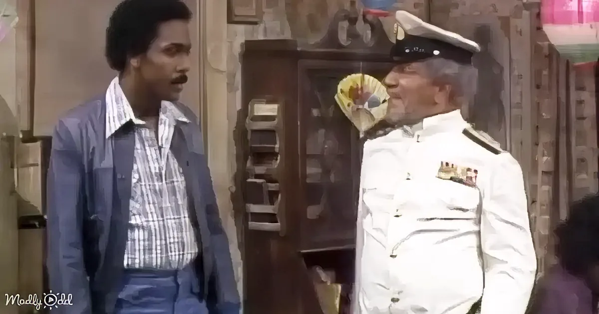 A man in a navy uniform is speaking to another man in a plaid shirt, both in a room with furniture and decorations.