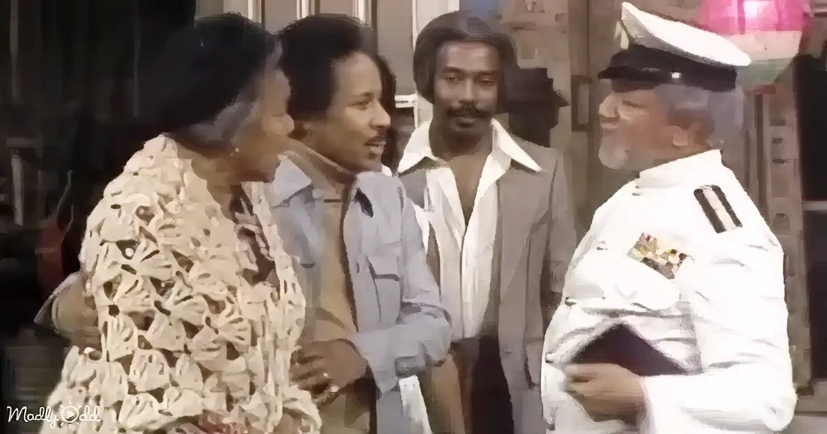 A man in a white military uniform is talking to a group of people, including a woman in a patterned coat.