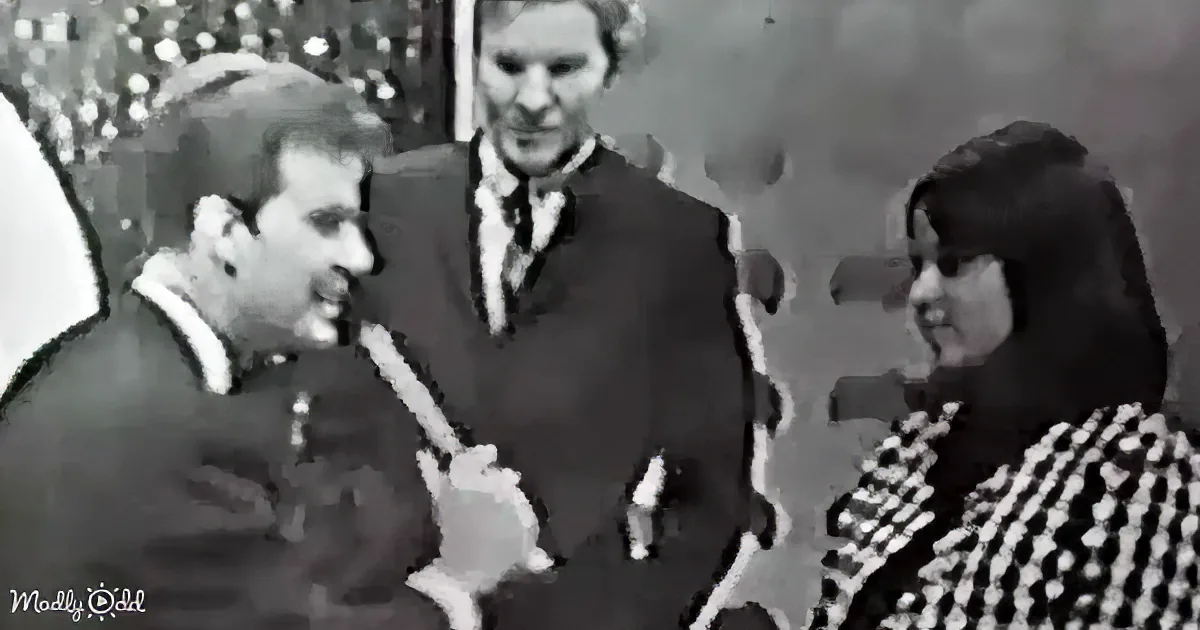 A man in a suit is holding a microphone, speaking to a woman in a checkered dress, with another person in the background.