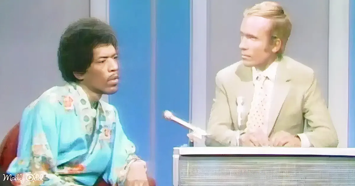 A man in a colorful shirt sits next to a man in a suit at a desk with a microphone.