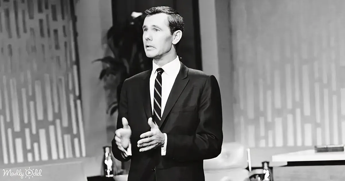 A man in a suit and tie stands on a stage, gesturing with his hands.