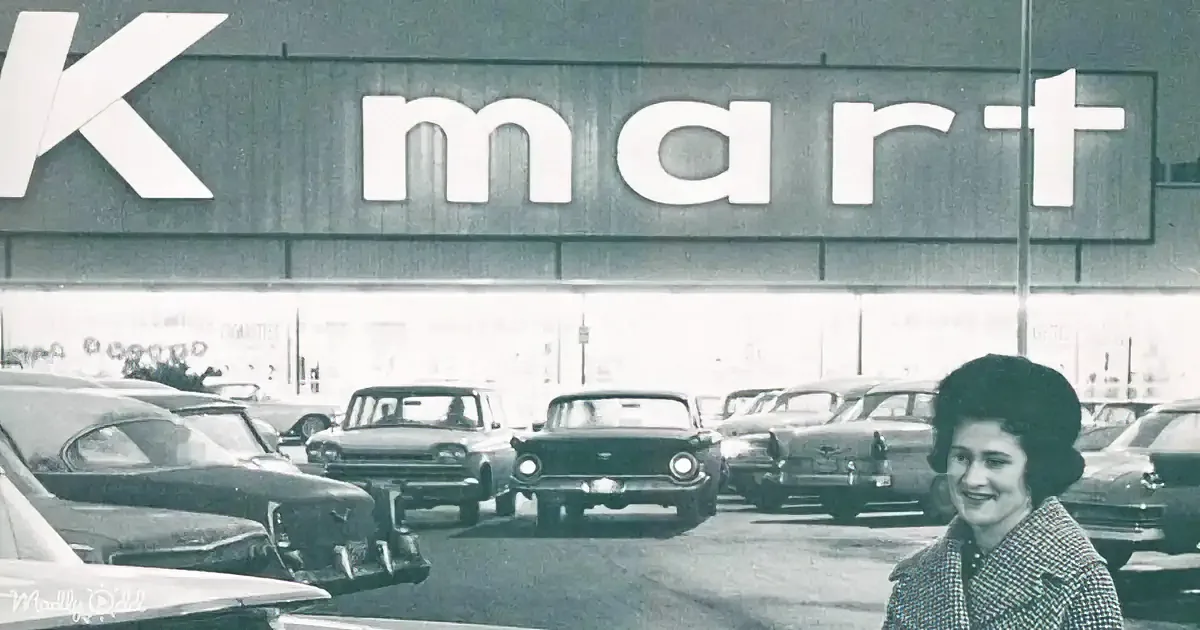 A woman smiles in a parking lot filled with vintage cars in front of a Kmart sign.