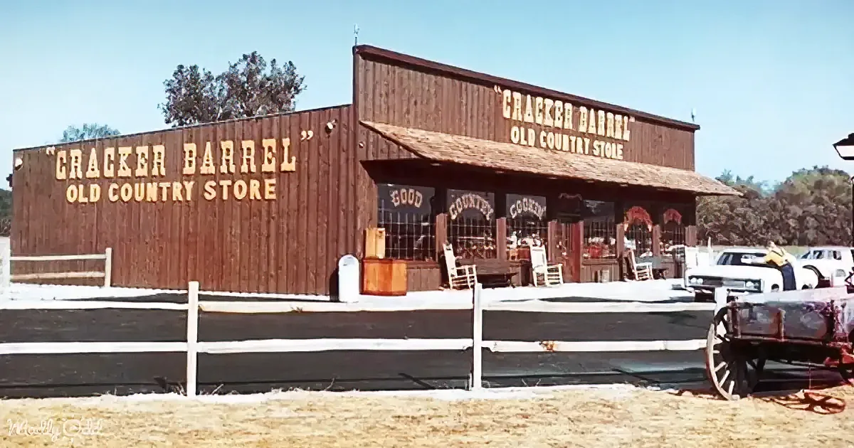 A Cracker Barrel Old Country Store with wooden facade, white fence, and vintage cars parked outside.