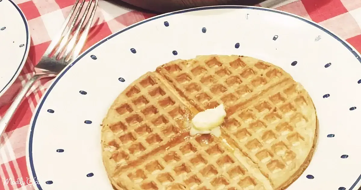 A golden waffle with butter sits on a white polka-dotted plate atop a red-and-white checkered tablecloth.