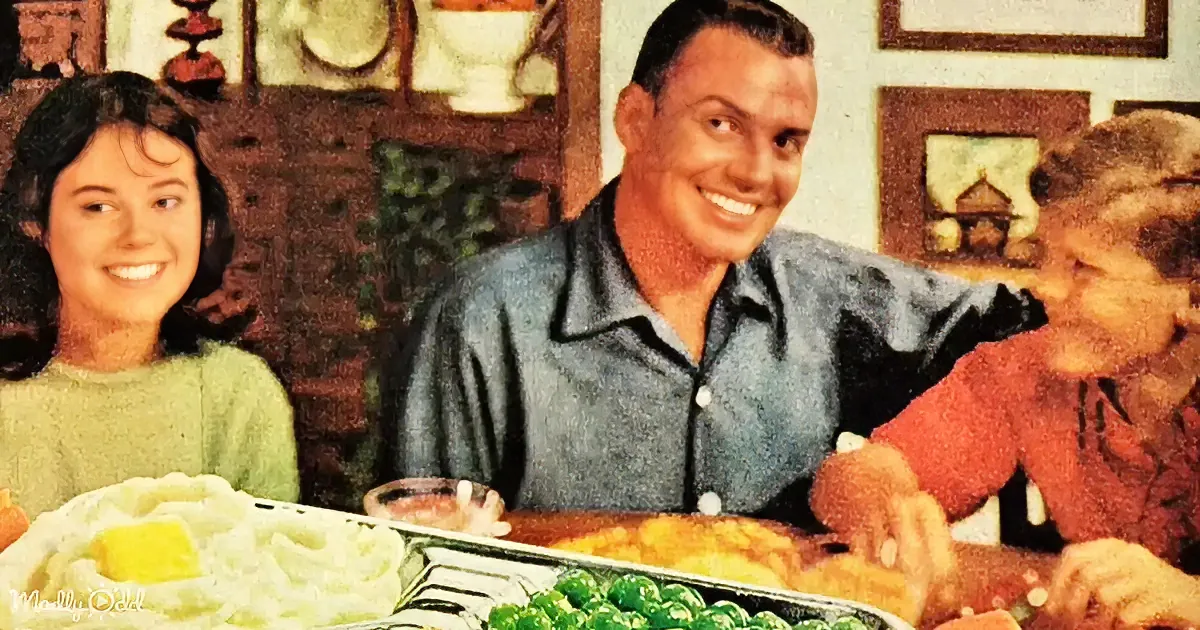 A smiling man sits at a table with a family, enjoying a meal of mashed potatoes, green beans, and other dishes.