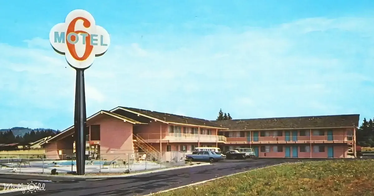 A Motel 6 sign stands tall beside a pink motel building with cars parked in front under a clear sky.