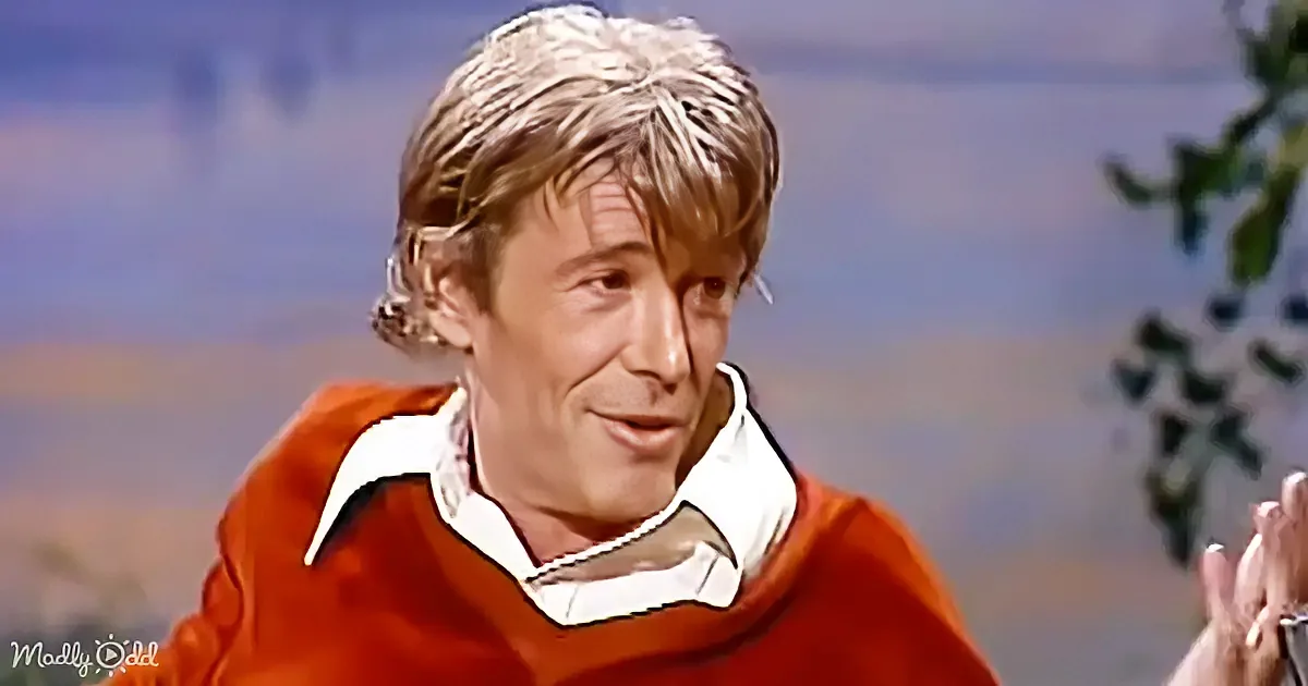 A man with blonde hair, wearing a red sweater and white shirt, smiles while sitting in front of a blurred background.