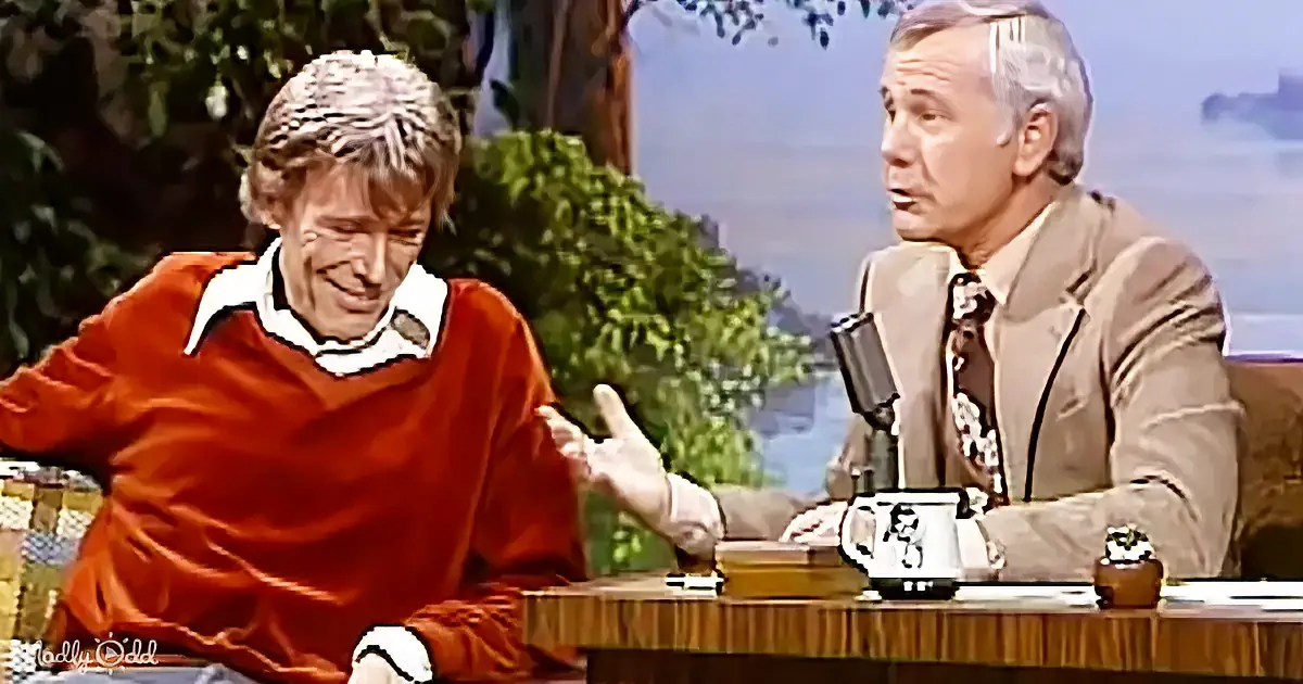 A man in a red sweater gestures animatedly while seated at a table with a host in a suit on a talk show set.
