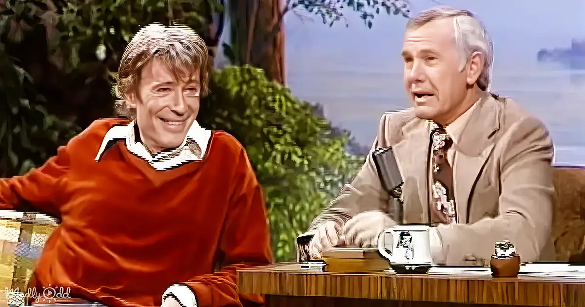 A man in a red sweater smiles while seated next to another man in a beige suit on a talk show set.