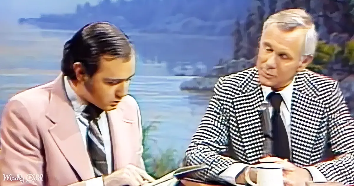 A man in a pink suit sits at a desk, looking down, while another man in a checkered suit sits beside him, holding a mug.