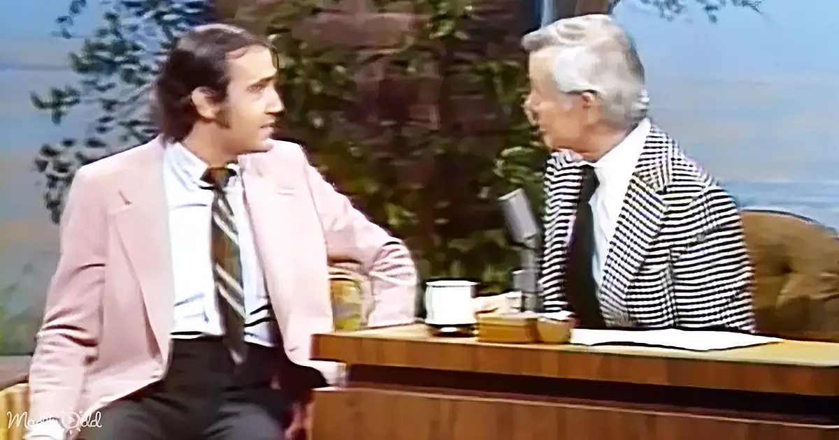 A man in a pink suit sits on a couch, engaged in conversation with another man in a striped suit on a talk show set.