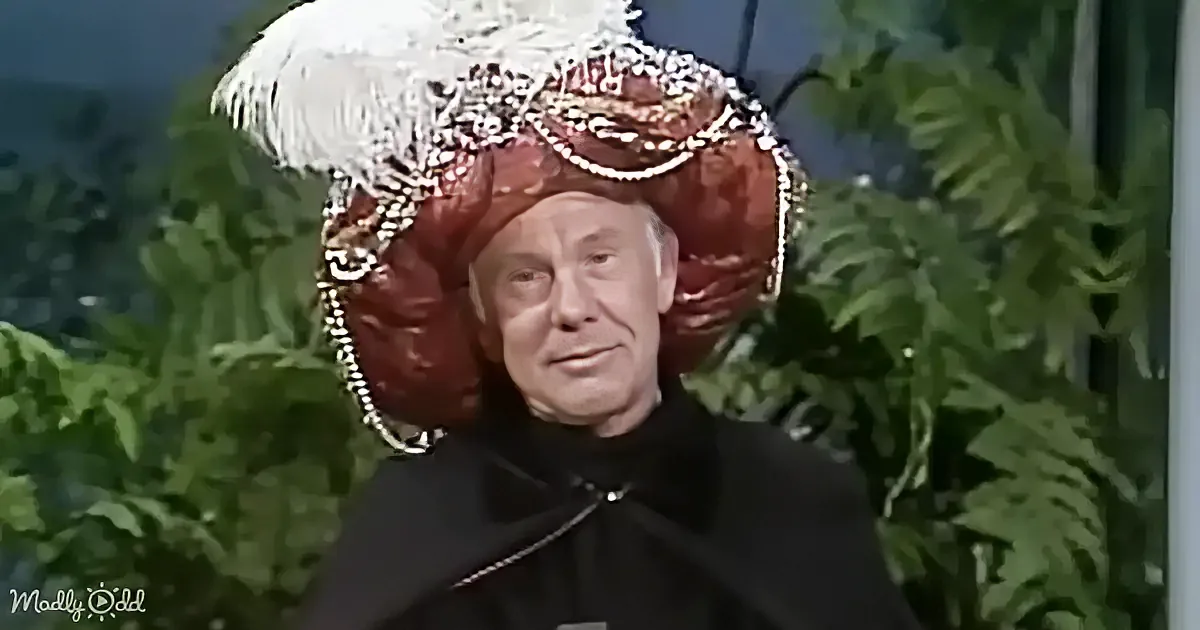 An elderly man in a black outfit and ornate red hat with white feathers stands against a green leafy background.