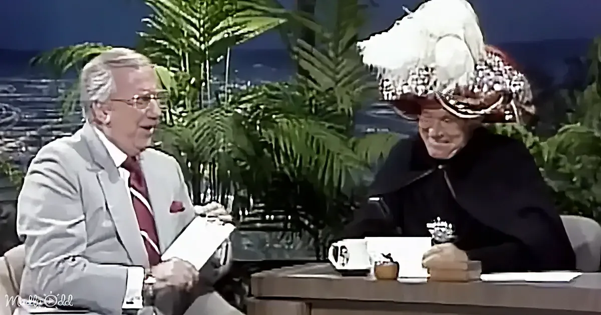 A man in a suit and tie is seated at a table with a person in a costume, both appearing to be on a talk show set.