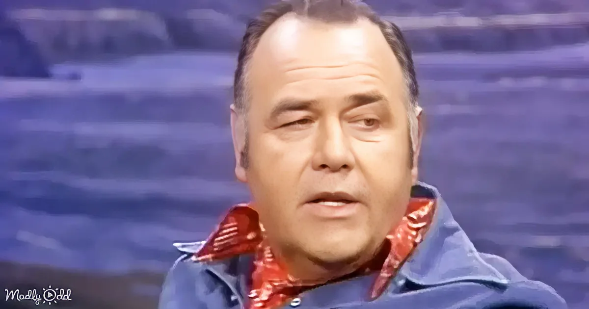 A man in a blue shirt and red bandana is speaking, with a blurred background.