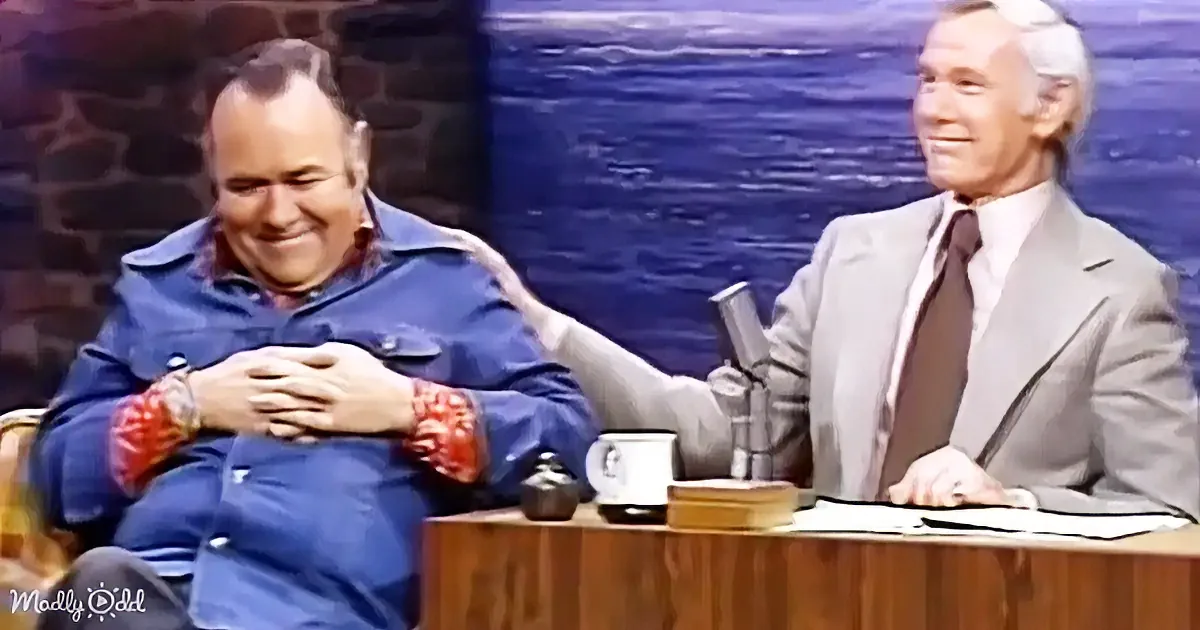 A man in a blue shirt sits with hands clasped, smiling, next to another man in a suit at a desk.