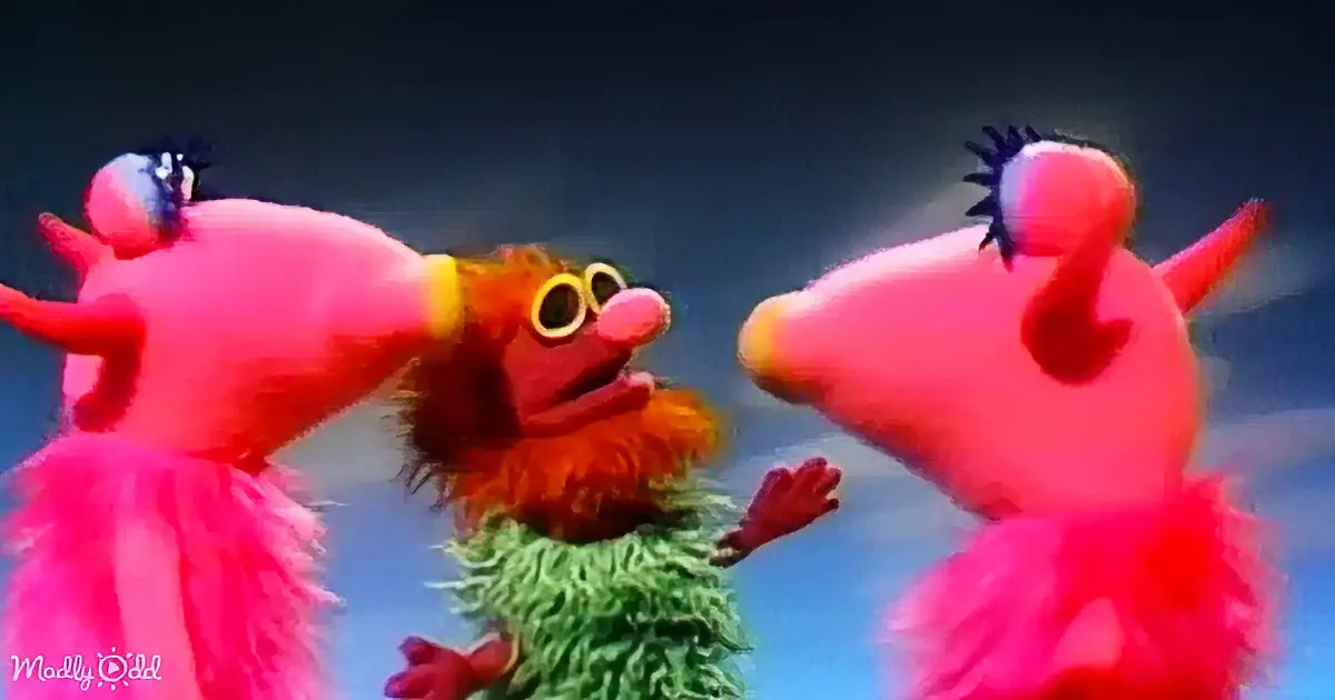 Three colorful, furry puppets with big eyes and red tongues stand together against a blue sky backdrop.