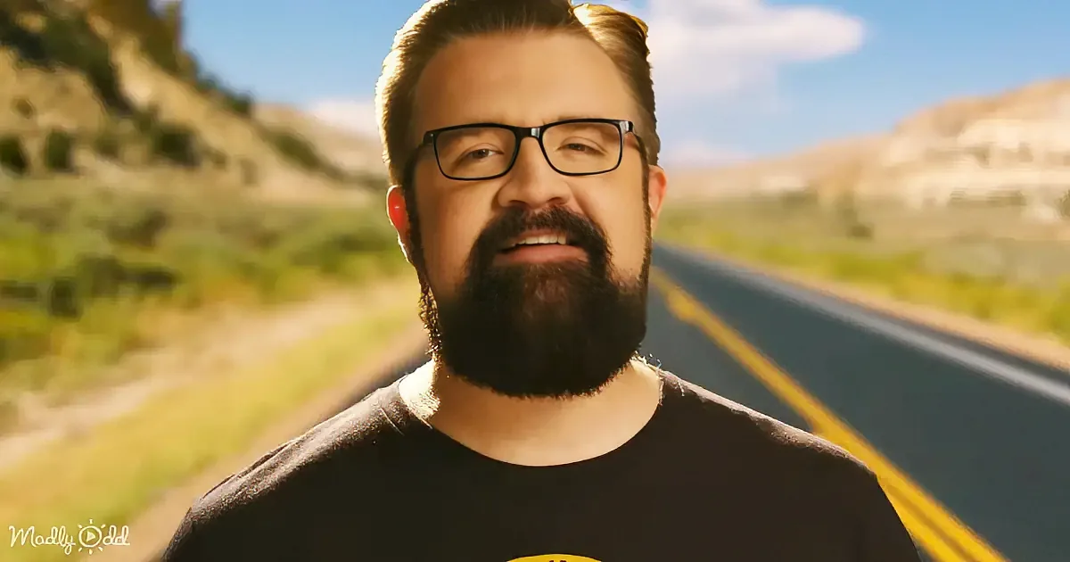 A man with a beard and glasses stands in front of a road with mountains in the background.