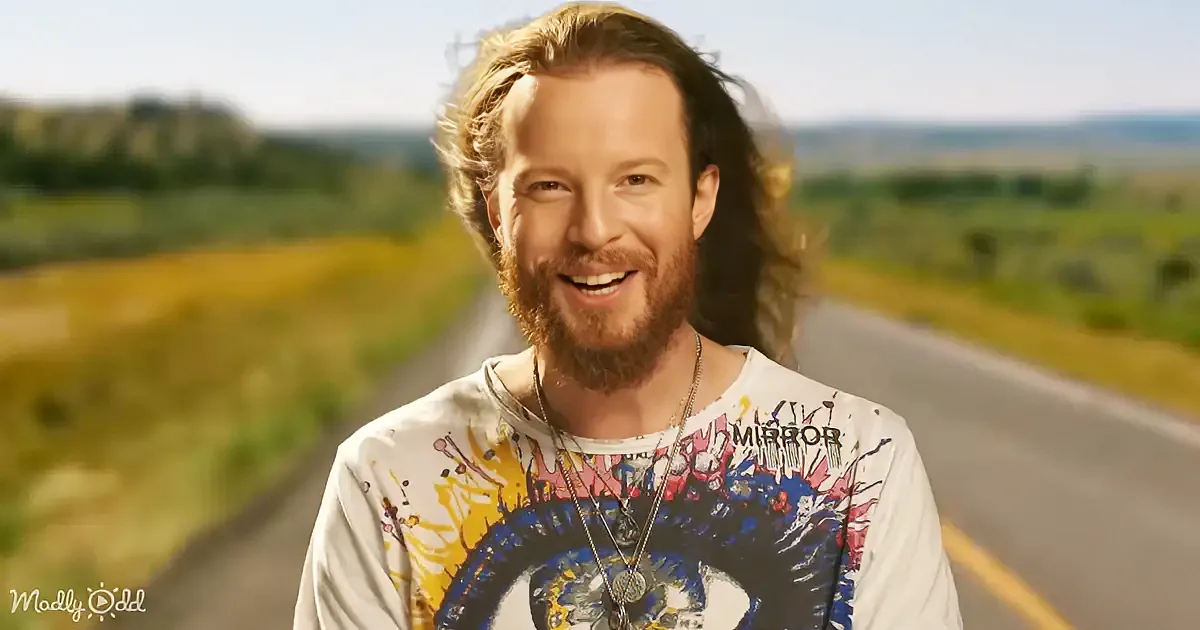 A man with long hair and a beard smiles in front of a road with a blurred landscape in the background.