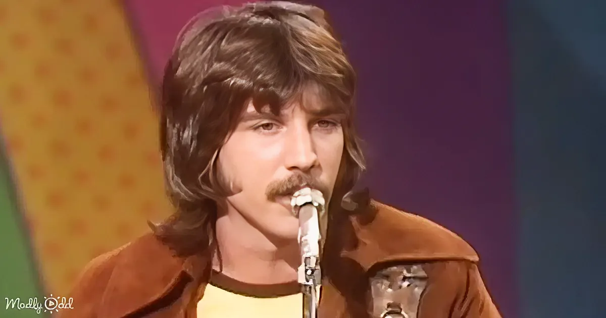 A man with long hair and a mustache sings into a microphone, wearing a brown jacket over a yellow shirt.