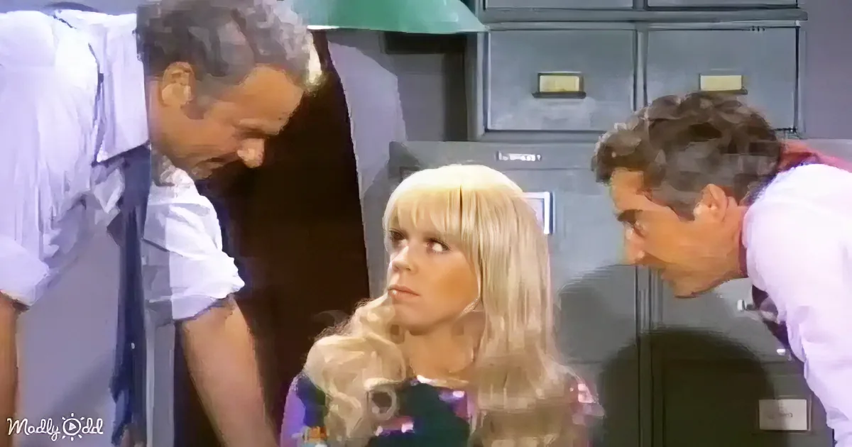Blonde woman with long hair sits between two men, one in a white shirt and the other in a purple shirt, in what appears...