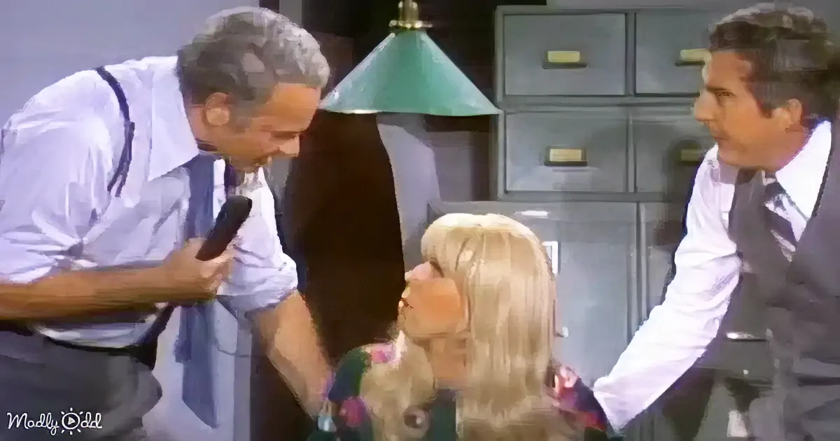 A man in a white shirt and suspenders holds a microphone, leaning towards a blonde woman in a floral dress, with another...