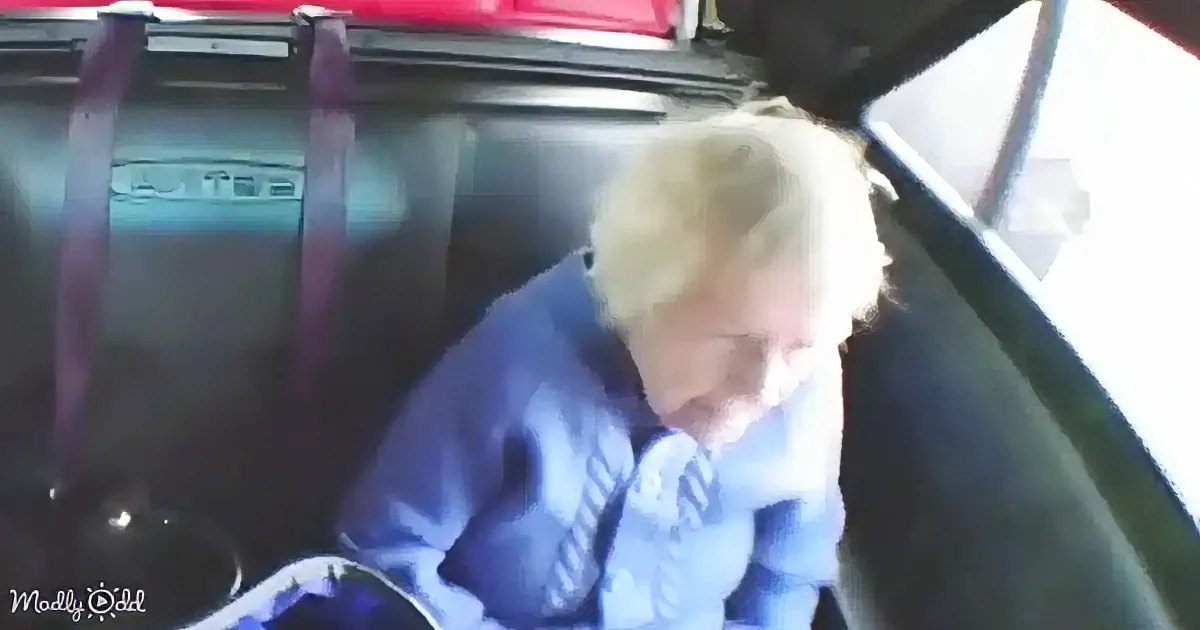 A person with blonde hair, wearing a blue shirt and tie, is sitting in a car, looking down.