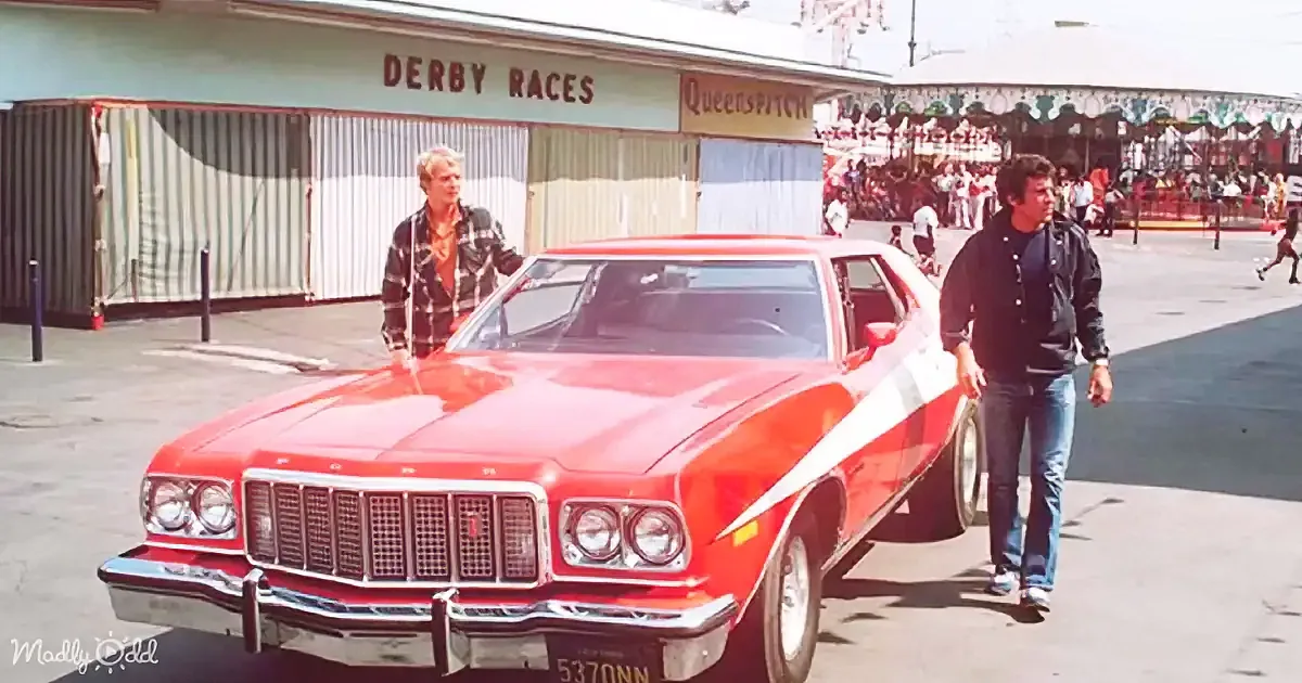 Two individuals walk beside a red Ford car in front of a "Derby Races" sign at a fairground.