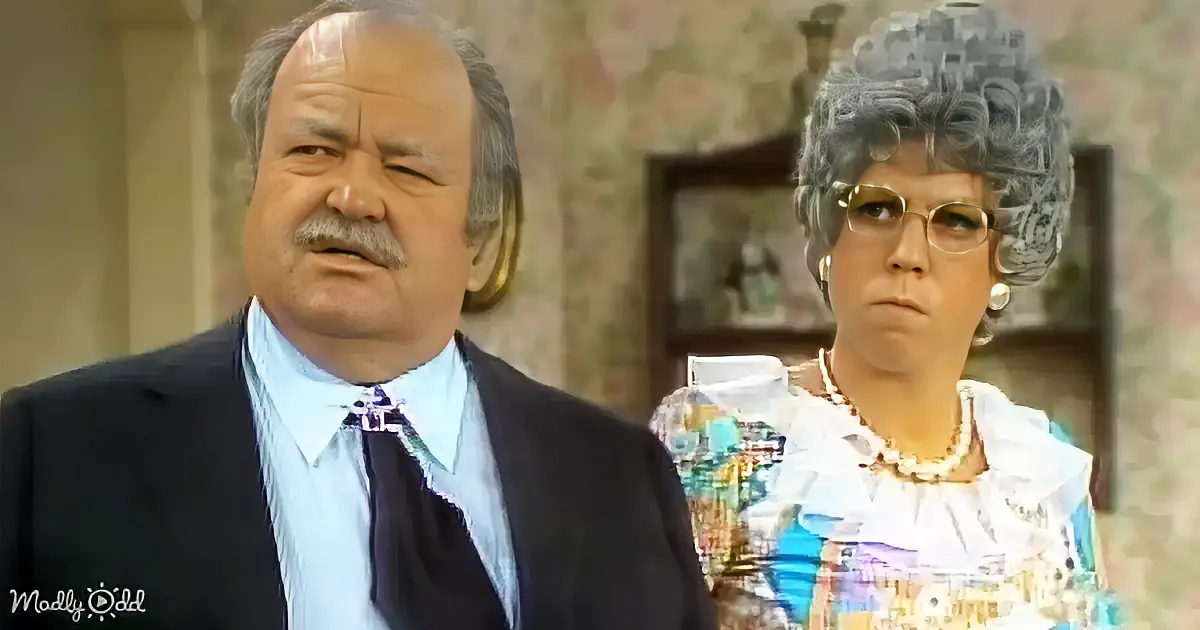A man in a suit and a woman in a colorful dress with glasses are standing indoors.
