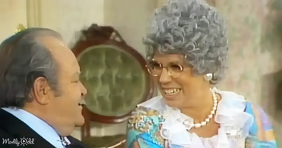 A smiling woman with a grey afro wig and pearls interacts with a man in a suit, both seated indoors.