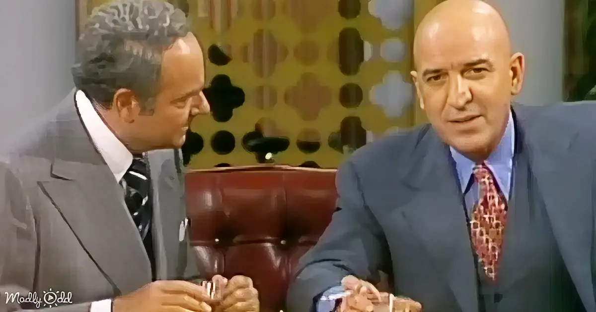 Two men in suits are seated, one holding a glass, against a patterned background.